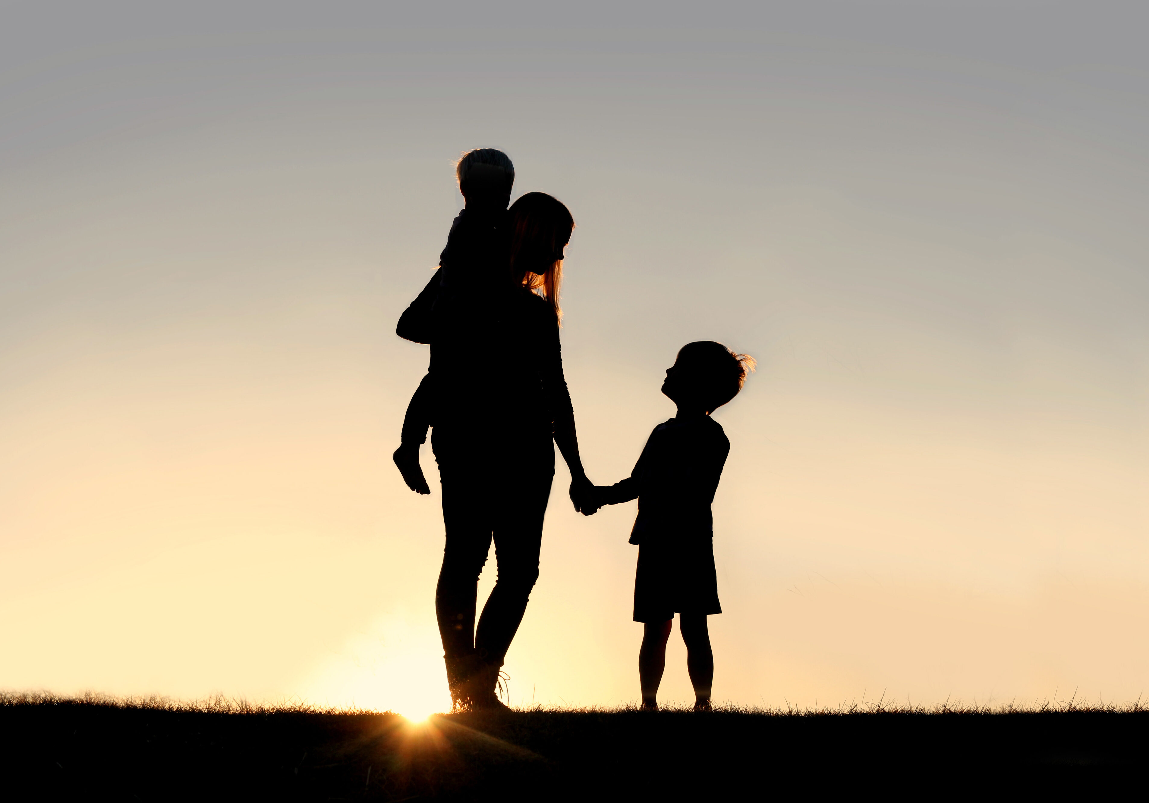 Silhouette of a young mother lovingly holding hands with her happy little child, while holding his baby brother, outside in front of a sunset in the sky.